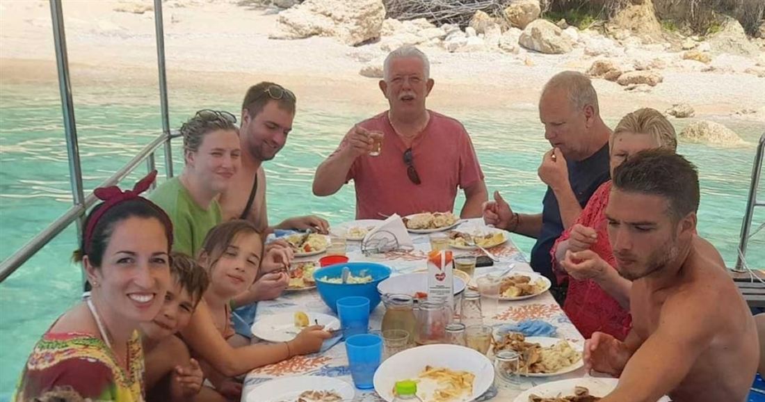 A family sharing a seafood lunch at a table on deck, anchored in clear water off a beach, on a Zakynthos fishing trip.