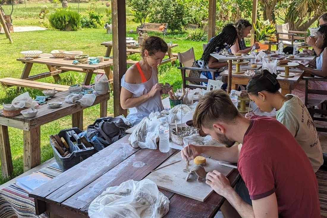 Guests shaping clay at wooden tables on a shaded veranda in a garden during a Zakynthos pottery class.