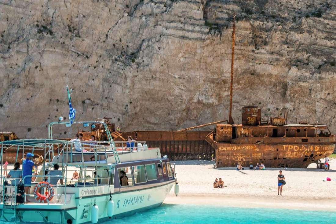 Tour boat anchored beside the rusted shipwreck on the white sand of Navagio beach below a tall limestone cliff