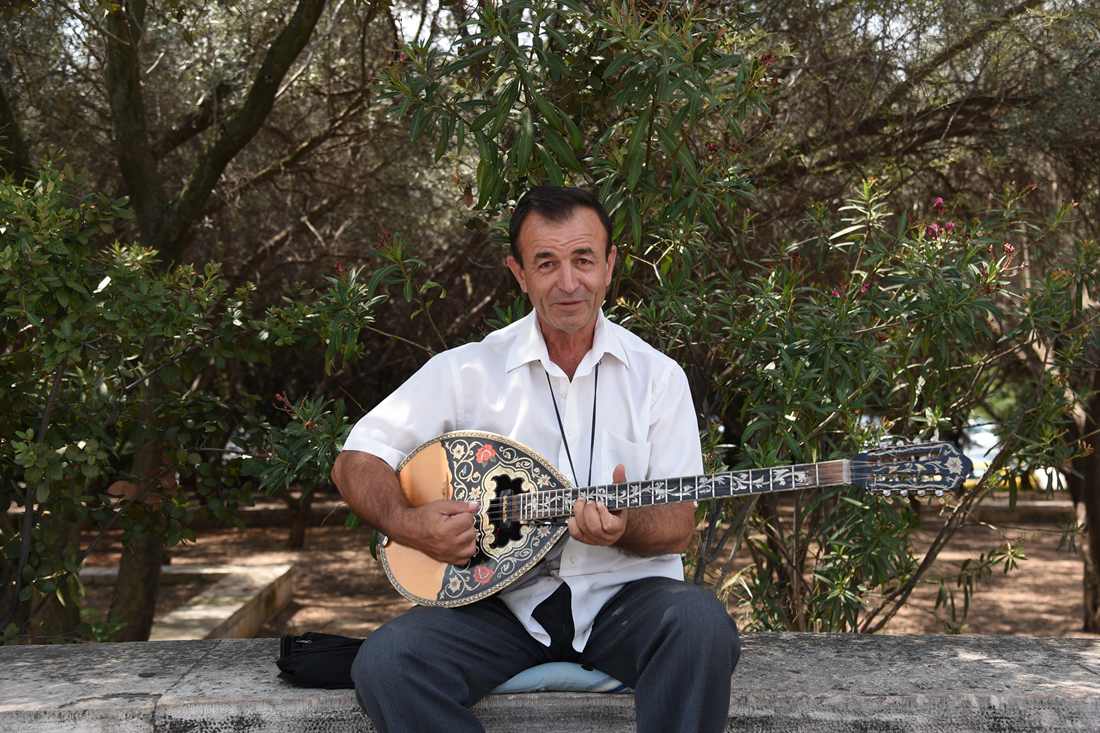 A bouzouki player in a white shirt sitting in a park and playing a bouzouki, with trees behind him